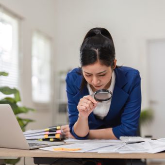 A person in a blue blazer leans over a desk, examining documents with a magnifying glass beside an open laptop; scattered highlighters and papers.