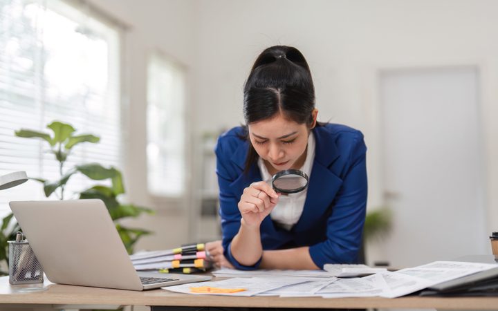A person in a blue blazer leans over a desk, examining documents with a magnifying glass beside an open laptop; scattered highlighters and papers.