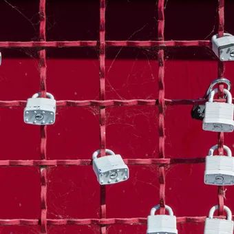 Rete di metallo rossa con lucchetti argentati fissati, sfondo rosso scuro, ragnatele sparse tra le griglie.