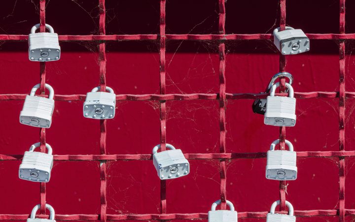 Rete di metallo rossa con lucchetti argentati fissati, sfondo rosso scuro, ragnatele sparse tra le griglie.