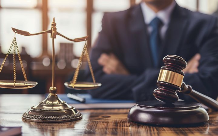 A judge's gavel and a set of scales sit on a wooden desk, with a person in a suit blurred in the background, implying a legal or courtroom setting.