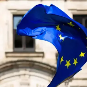 A torn European Union flag waving in front of a historic stone building with large windows.