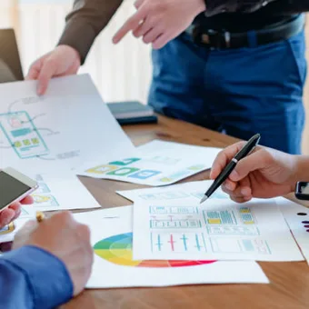 People sharing and discussing colourful charts and documents around a wooden table, with laptops and smartphones in a bright office setting.