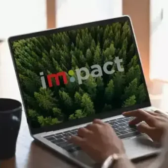 A person using a laptop displays the word "impact" with a forest background, on a wooden table with a cup and a container of pencils nearby.