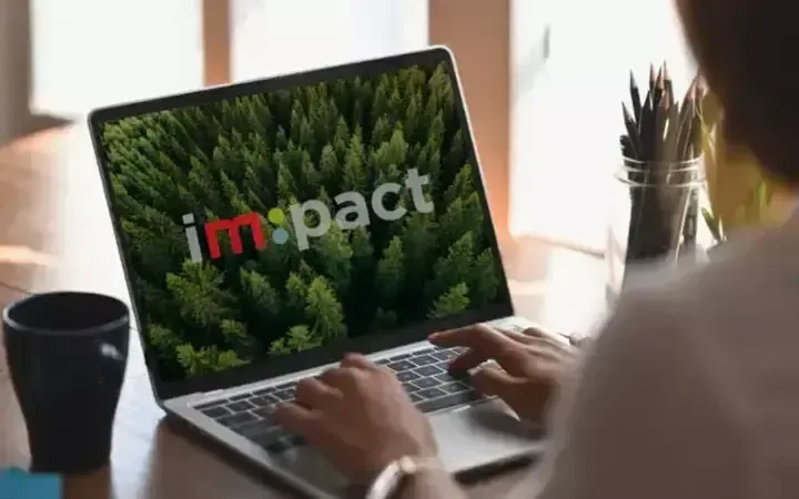 Person using a laptop with a forest background on the screen displaying the word "impact". The person is typing, and there is a cup and pencil holder nearby.
