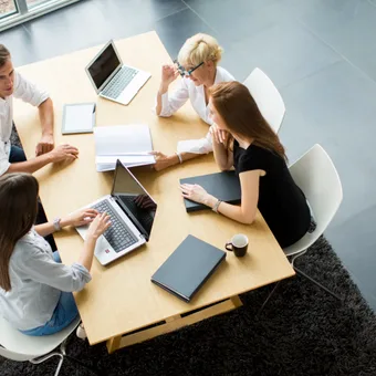 Five people sit around a light wooden table with laptops, notebooks, and a coffee mug, engaged in a discussion in a modern office setting.
