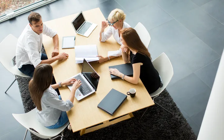 Five people sit around a light wooden table with laptops, notebooks, and a coffee mug, engaged in a discussion in a modern office setting.