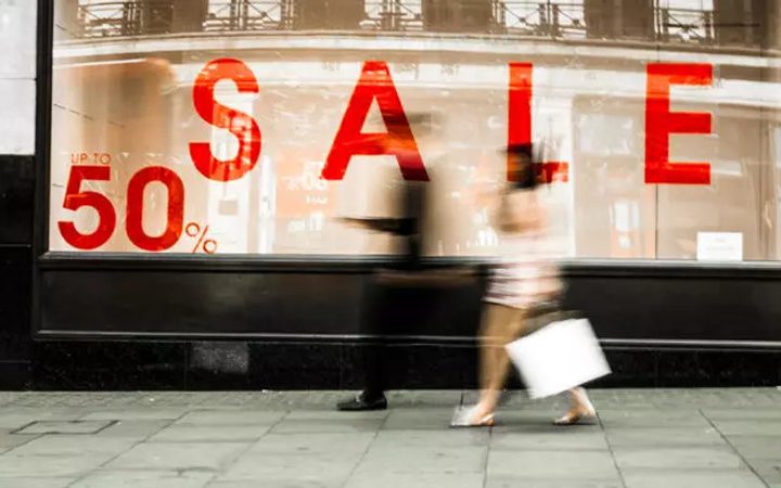 Blurred pedestrians walking past a store window with large red "SALE" letters and a sign saying "up to 50%" discount.