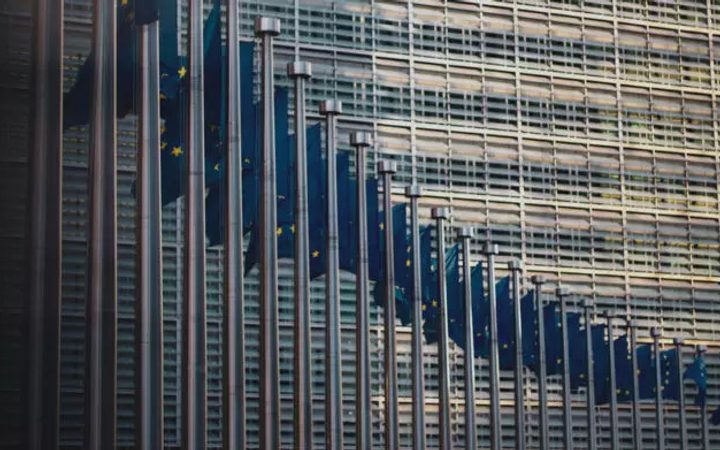 Multiple European Union flags hanging vertically in front of a modern glass and metal office building.