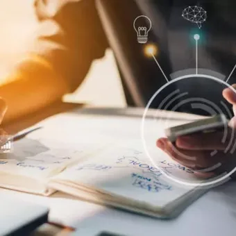 Person using a smartphone and writing in a notebook on a desk with digital icons and graphs overlaid.