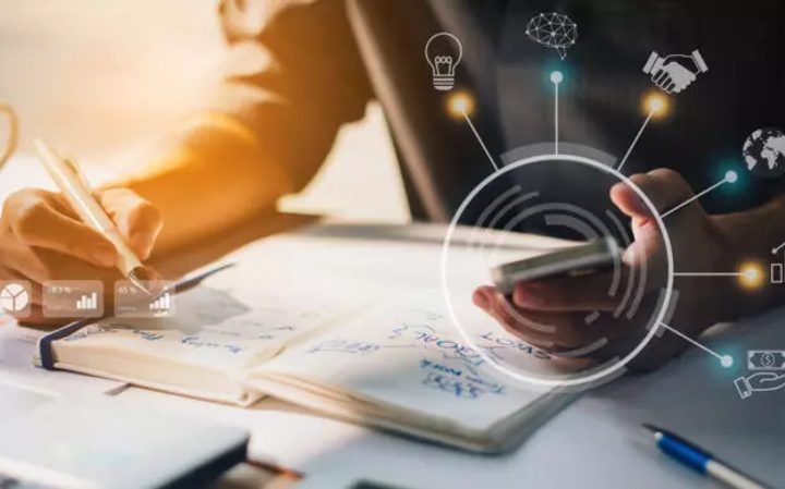 Person using a smartphone and writing in a notebook on a desk with digital icons and graphs overlaid.