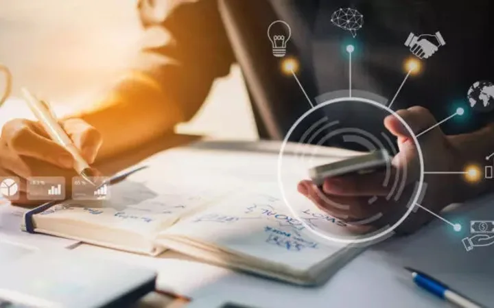 Person using a smartphone and writing in a notebook on a desk with digital icons and graphs overlaid.