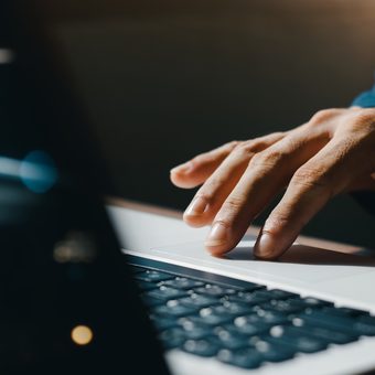 Close-up of a hand typing on a laptop keyboard; blue sleeve visible, with a softly lit, blurred background and bokeh lights.