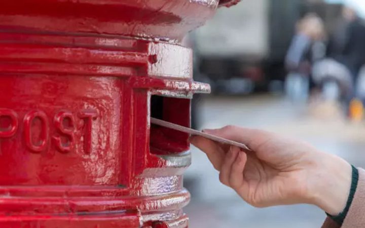 A hand posts a letter into a red British post box with the word "POST" embossed on its front. The background is blurred with people and vehicles.