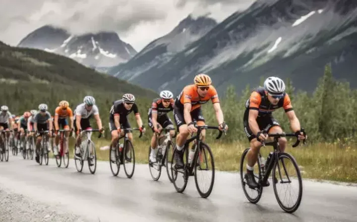 Group of cyclists racing on a mountain road with snow-capped peaks and green forested slopes in the background.