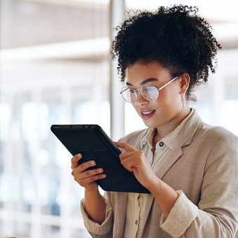 Person with curly hair and round glasses, wearing a beige blazer, looks at and interacts with a tablet in a bright indoor space.
