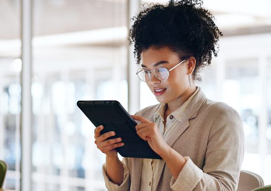 Personne aux cheveux bouclés et lunettes, vêtue d’un blazer beige et d’une chemise claire, tient une tablette numérique dans un espace lumineux.