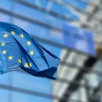 European Union flag with yellow stars on a blue background flying in front of a modern glass building.