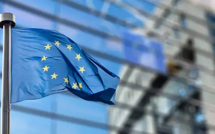 European Union flag with yellow stars on a blue background flying in front of a modern glass building.