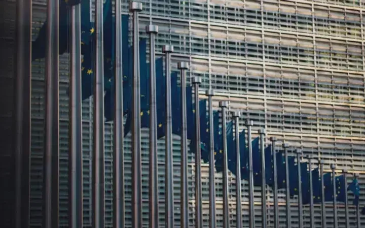 Multiple EU flags hang in front of a modern glass building, reflecting the sky and surrounding structures. The flags are aligned along tall flagpoles.