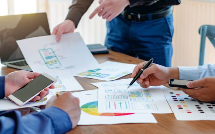People working together at a table with charts, graphs, and electronic devices, discussing and collaborating on a project or presentation.
