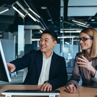 Two professionals are engaged in a discussion in a modern office, with the man pointing at a computer monitor.