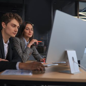 Two young professionals work together at a desk in a modern office, focused on a computer screen, with papers and pens on the desk.