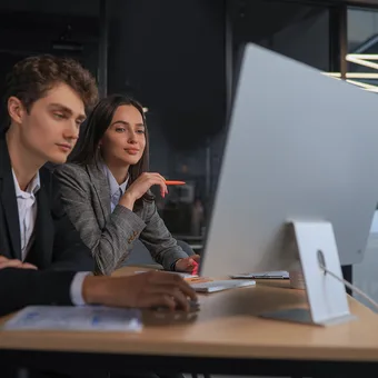 Two young professionals work together at a desk in a modern office, focused on a computer screen, with papers and pens on the desk.