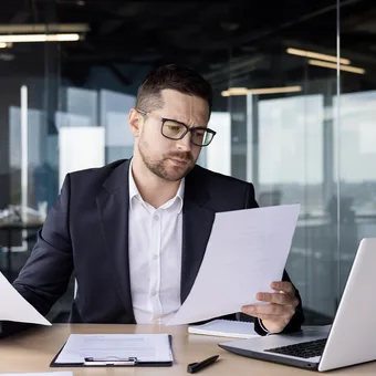 Person in a suit with glasses sitting at a desk, looking at papers, with a laptop and documents, in a modern office setting.