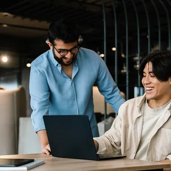 Two people sharing a laugh while looking at a laptop, with notebooks and a tablet on the table in a modern, well-lit office space.