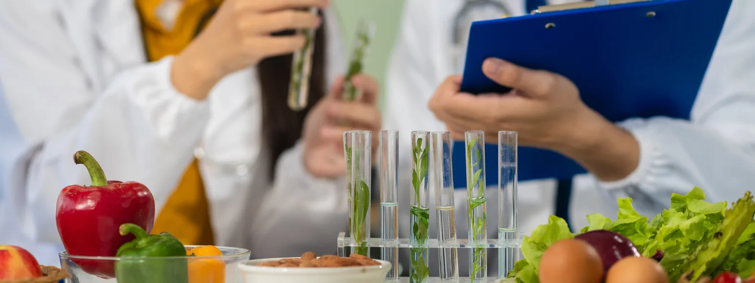 Scientists examining plant samples with test tubes and a clipboard surrounded by vegetables and bowls on a table.