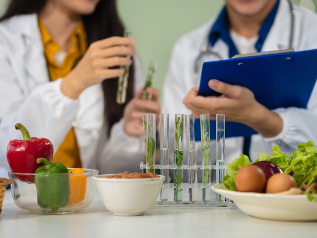 Scientists examining plant samples with test tubes and a clipboard surrounded by vegetables and bowls on a table.