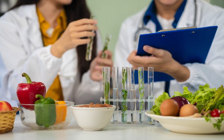 Scientists examining plant samples with test tubes and a clipboard surrounded by vegetables and bowls on a table.