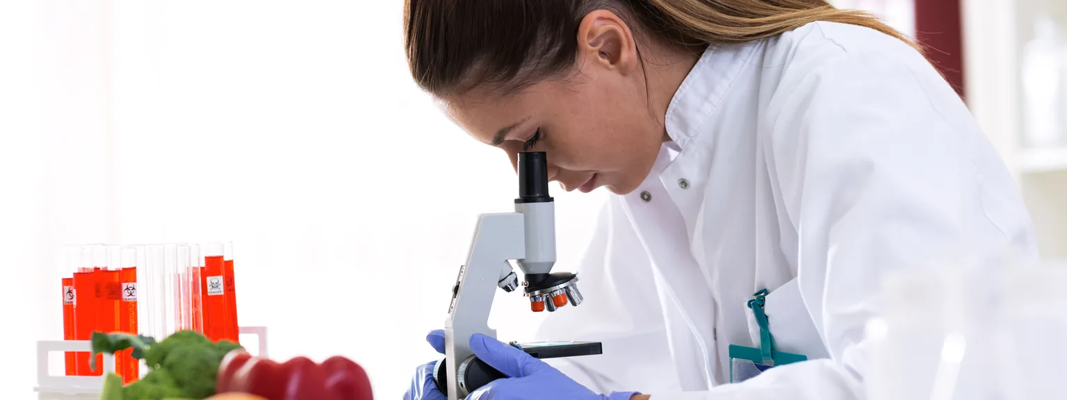 Scientist examining produce with a microscope, surrounded by fruits and lab equipment in a laboratory setting.