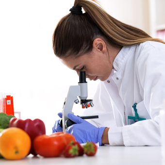 Scientist examining produce with a microscope, surrounded by fruits and lab equipment in a laboratory setting.