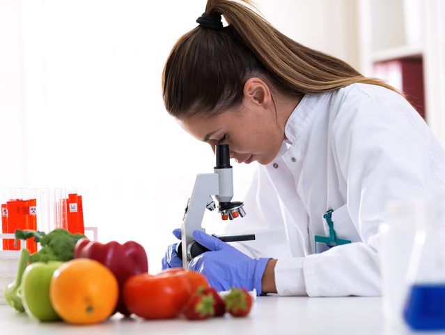 Scientist examining produce with a microscope, surrounded by fruits and lab equipment in a laboratory setting.