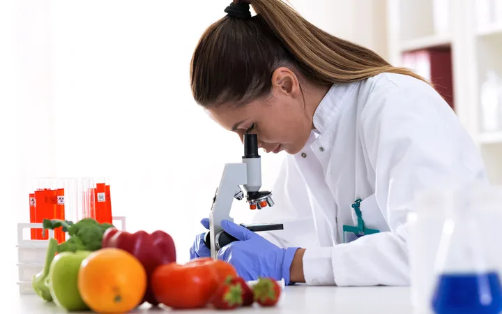 Female scientist examining produce with a microscope, surrounded by fruits and lab equipment in a laboratory setting.