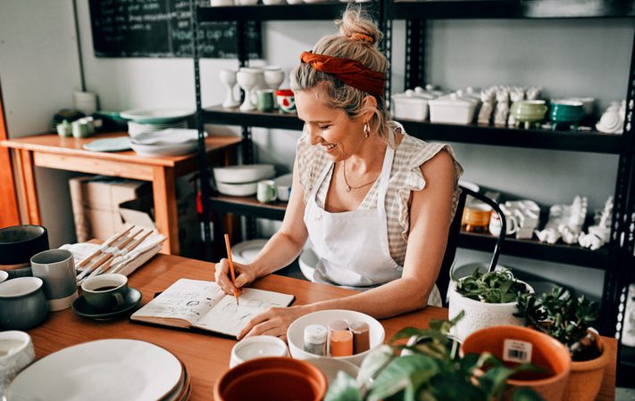 Person smiling while drawing in a notebook at a pottery studio surrounded by ceramic items and plants.