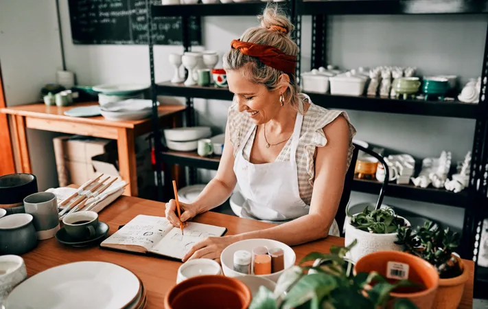 Person smiling while drawing in a notebook at a pottery studio surrounded by ceramic items and plants.