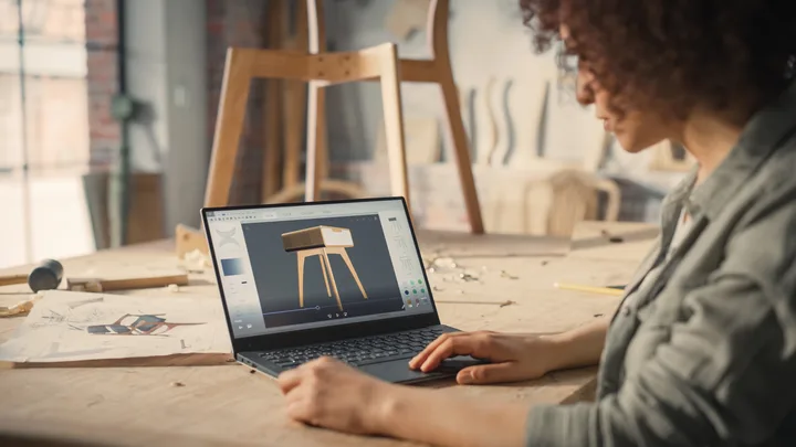 A person with curly hair designs a wooden stool with a laptop in a creative workspace, surrounded by sketches and tools on a wooden table.