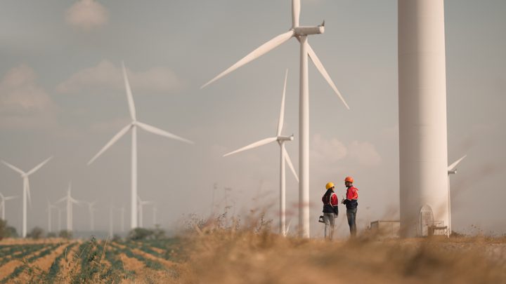 Two workers in safety helmets and vests converse near wind turbines in a field under a cloudy sky.