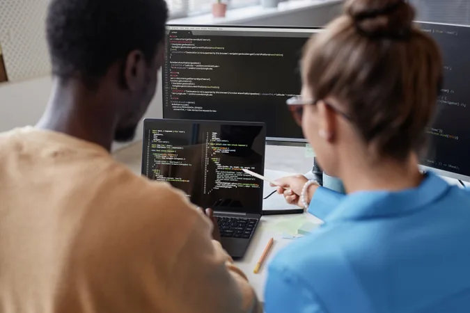 Two people collaborate on coding, with one pointing at a laptop displaying programming code, while their screens show more code in the background.