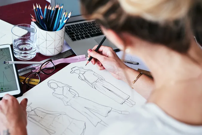 A person sketches fashion designs on paper surrounded by coloured pencils, glasses, a ruler, a smartphone, and a laptop on a worktable.