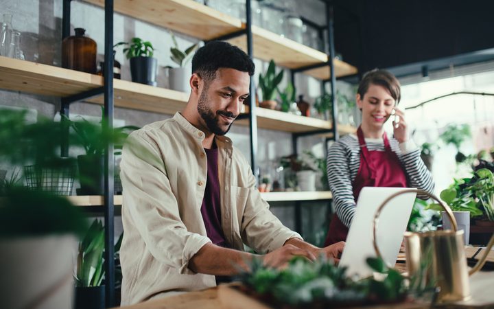 Person working on a laptop in a plant shop with shelves of potted plants and another person on the phone in the background, both smiling.