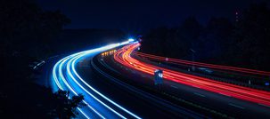 Light trails of blue and red moving along a winding highway at night, with dark trees on either side and a starry sky overhead.
