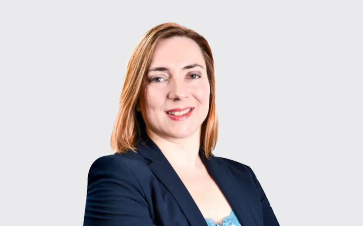 A person with shoulder-length light brown hair, smiling confidently, wearing a navy blazer against a light grey background.