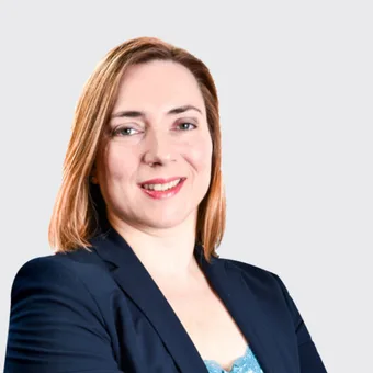 A confident person with shoulder-length auburn hair, wearing a navy blazer, smiling against a light grey background.