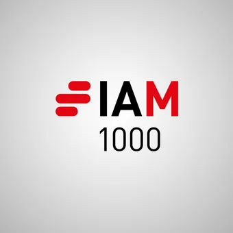 Portrait of a person in a navy blazer and white shirt, with short dark hair, against a grey gradient studio background.