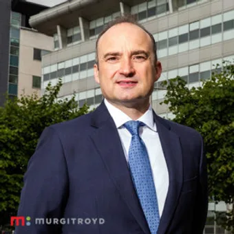 A person in a navy suit and blue tie standing outdoors in front of modern office buildings with trees, smiling at the camera.