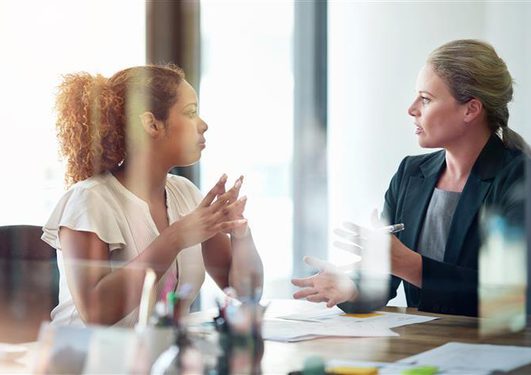 Two professionals converse at a desk in an office; one with curly hair in a white blouse, the other in a blazer, with papers on the table.